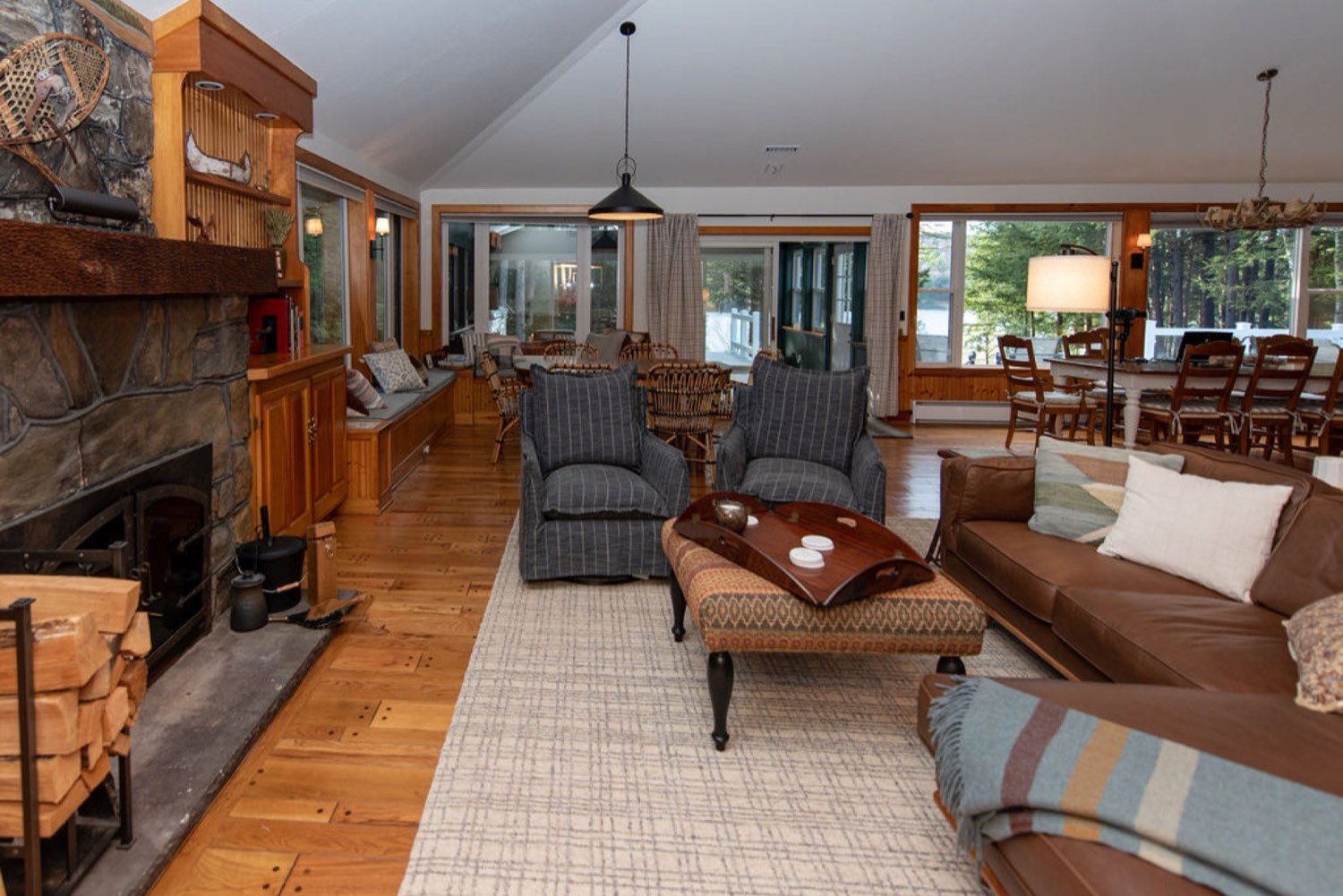 The great room at Camp Sunnyside with a fieldstone hearth stacked with firewood, leather sofas, and a wall of glass toward the lake.